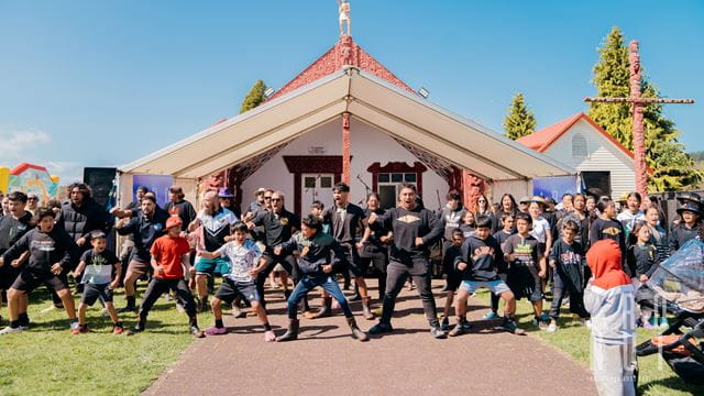 A group of children and adults perform a haka outside a meeting house on a sunny day.