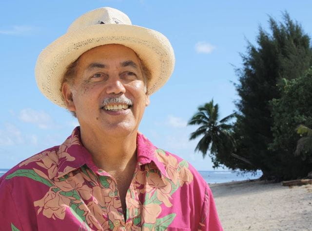 Dr Jon Jonassen, an older Cook Island man, smiles kindly, looking off camera, standing on a beautiful beach wearing a pink Island shirt and a beige hat