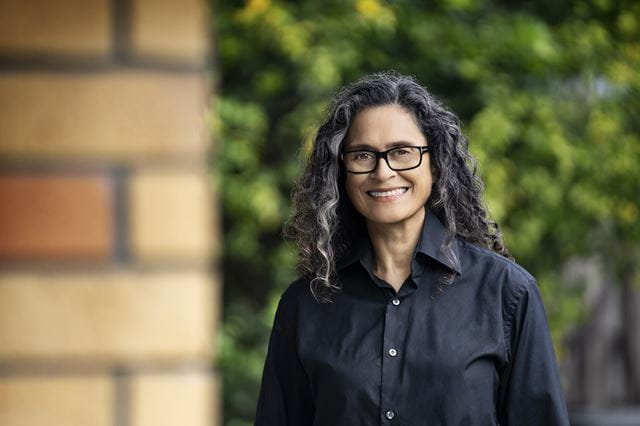 Gina Cole wears a black shirt and has shoulder-length curly hair. She wears glasses and smiles at the camera in front of a green background.