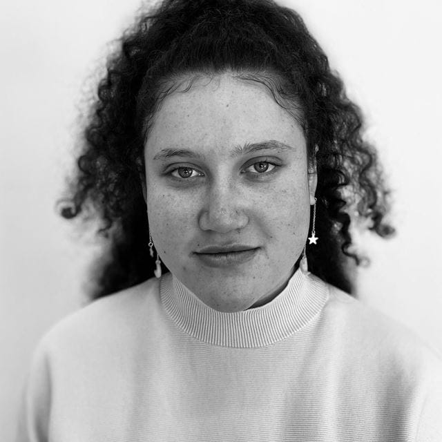 Teherenui Koteka, a young Cook Island woman, looks directly at the camera with a slight smile. She is wearing a high-neck white jumper and has her curly hair in a high ponytail.