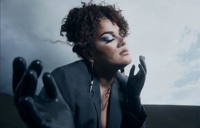 A moody, glamourous shot of Lady Shaka, a Pasifika woman, wearing gloves and a suit with blue eyemakeup