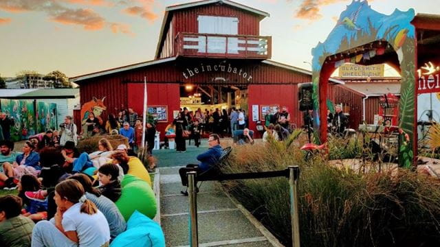 People gather and sit outside of a wooden building decorated with bunting on a rainbow floor design, within The Incubator village.